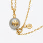 A gold chain necklace featuring a round gray pearl pendant with a gold T-Logo cross in the center, alongside a small gold brand tag, on a light gray background.
