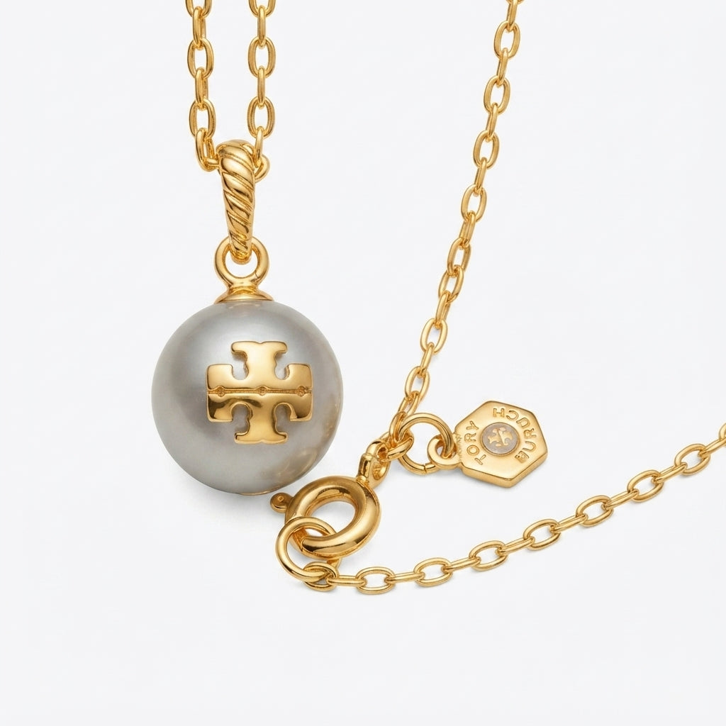 A gold chain necklace featuring a round gray pearl pendant with a gold T-Logo cross in the center, alongside a small gold brand tag, on a light gray background.