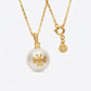 A gold chain necklace featuring a large white pearl pendant with a gold cross motif and a small hexagonal charm engraved with 'TORY Burch', shown on a light gray background.