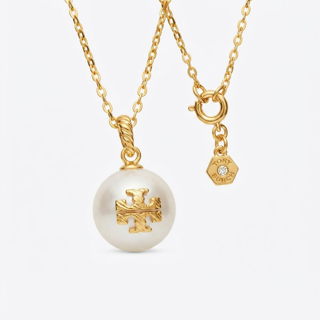 A gold chain necklace featuring a large white pearl pendant with a gold cross motif and a small hexagonal charm engraved with 'TORY Burch', shown on a light gray background.
