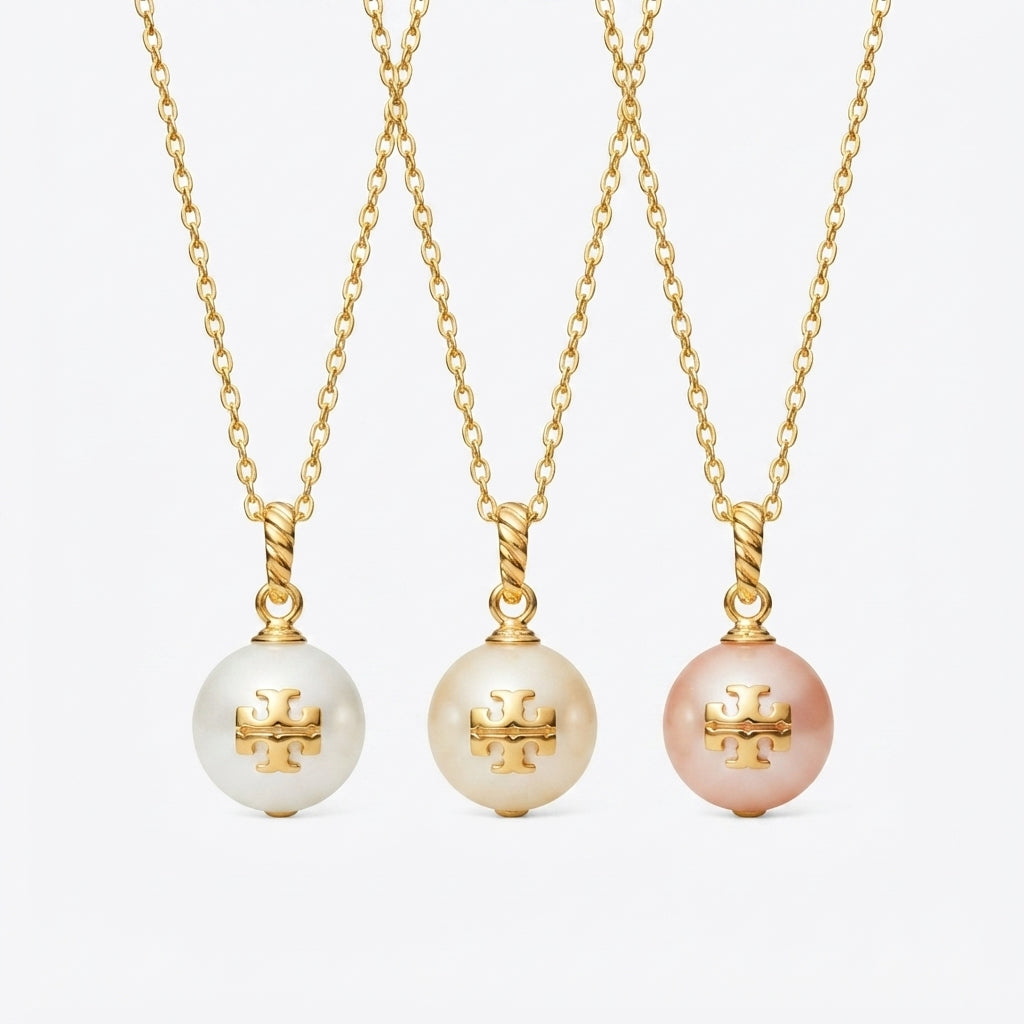 Three gold chain necklaces with round pearl-like pendants are shown against a light gray background. Each pendant features a gold monogram logo, with the left and center pendants in white/cream and the right pendant in soft pink.