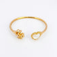 A gold-plated open cuff bangle bracelet featuring a textured flower on one end and a heart-shaped mother-of-pearl inlay on the other, set against a plain white background.