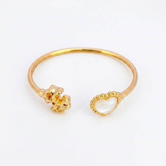 A gold-plated open cuff bangle bracelet featuring a textured flower on one end and a heart-shaped mother-of-pearl inlay on the other, set against a plain white background.