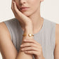 A close-up of a model wearing a delicate gold cuff bracelet with a heart-shaped pearl and a small gold flower accent on the other side, paired with a light gray ribbed sleeveless top.