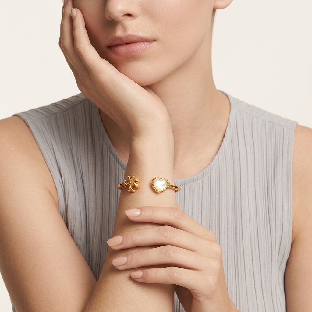 A close-up of a model wearing a delicate gold cuff bracelet with a heart-shaped pearl and a small gold flower accent on the other side, paired with a light gray ribbed sleeveless top.