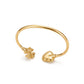 A gold-plated open cuff bangle bracelet featuring a small flower charm on one end and a heart-shaped bead border charm on the other, displayed on a clean white background.