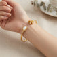 A close-up of a person's wrist wearing two gold-toned bangle bracelets. One bracelet features a smooth oval white pearl-like center, while the other has a textured gold floral or butterfly motif.
