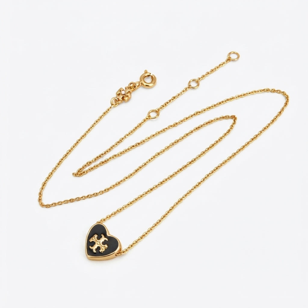 A delicate gold chain necklace featuring a small heart-shaped pendant with a black center and a gold monogram logo, displayed on a clean white background.