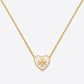 A gold heart-shaped pendant necklace with an open heart design and a central monogram logo, suspended from a delicate gold chain against a light gray background.