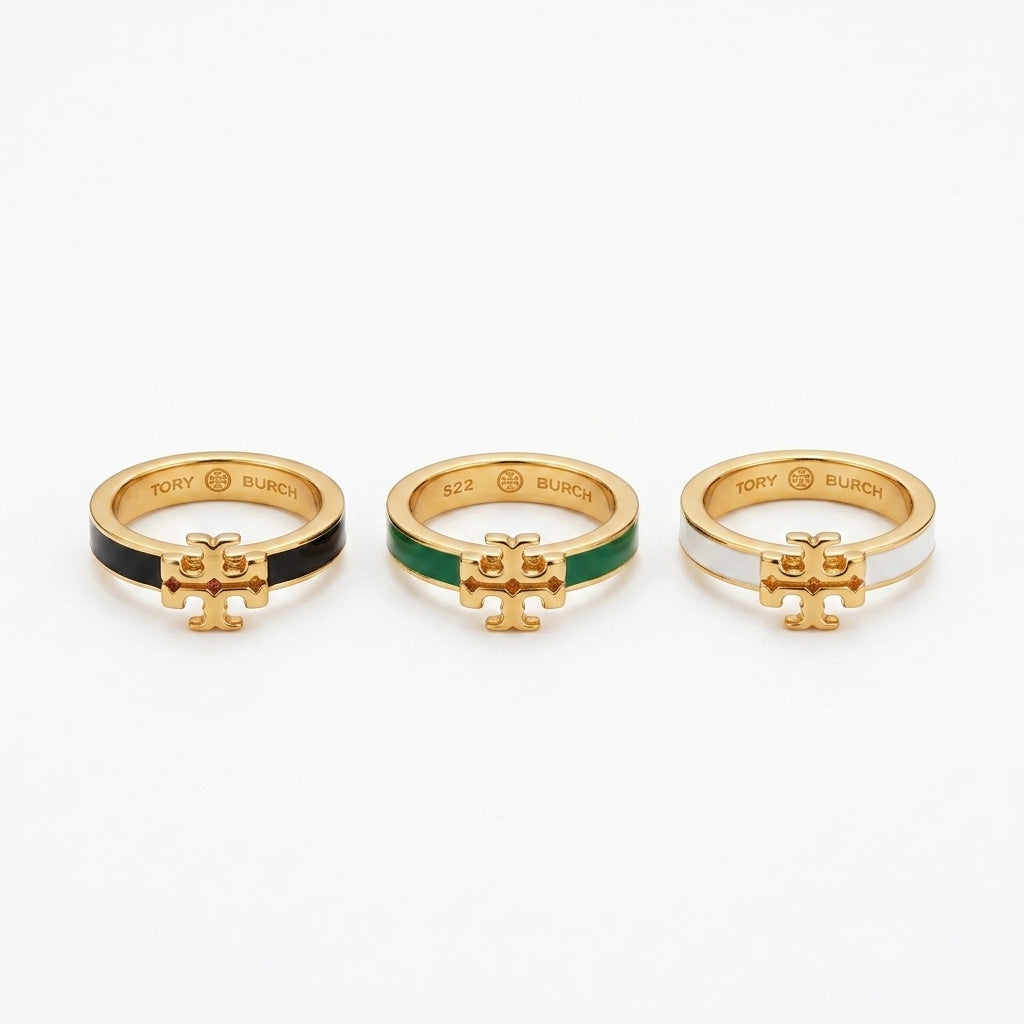 Three gold rings with the Tory Burch logo charm, shown side by side on a white background. The left ring has a black enamel section, the middle ring has a green enamel section, and the right ring has a white enamel section.
