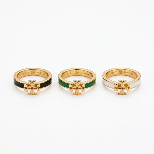Three gold rings with the Tory Burch logo charm, shown side by side on a white background. The left ring has a black enamel section, the middle ring has a green enamel section, and the right ring has a white enamel section.