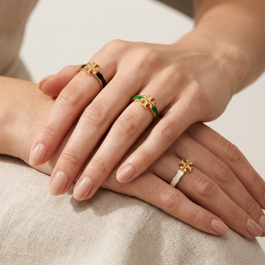 Three gold rings with green and white enamel details are shown on a person's hands, resting on a light beige fabric surface.