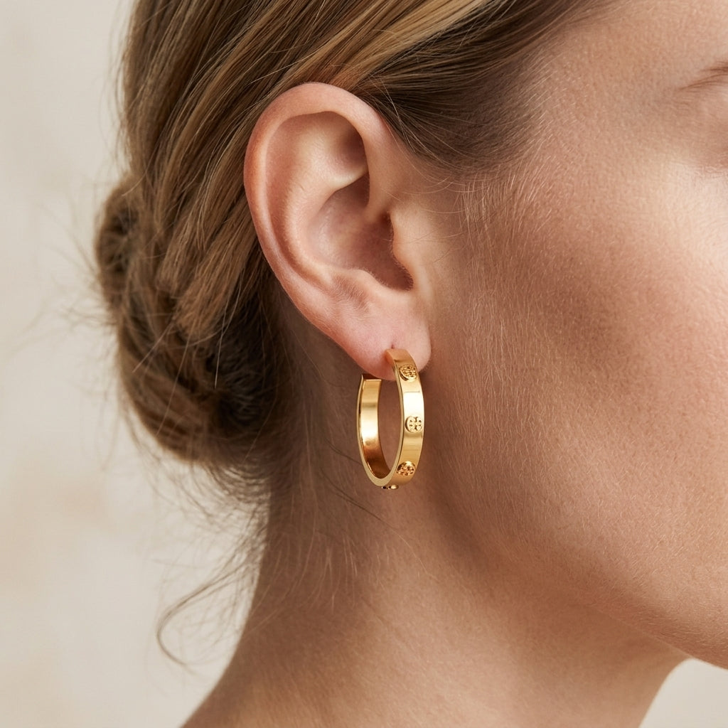 A close-up of a model's ear wearing a small gold hoop earring with engraved details, set against a soft beige background.