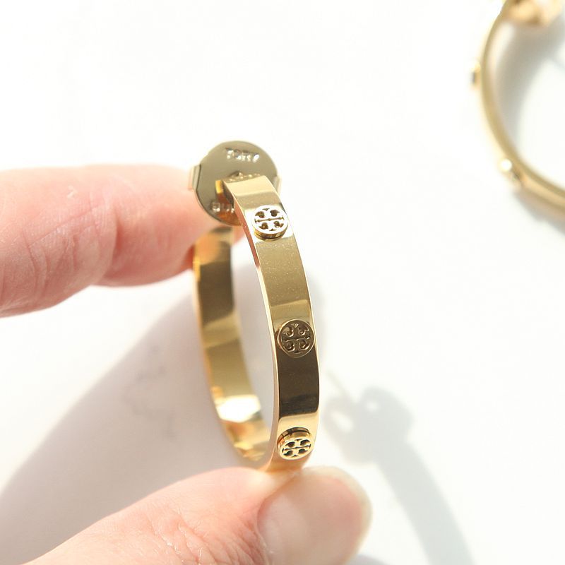 A gold bangle bracelet with circular logo details, held by a hand against a white background.