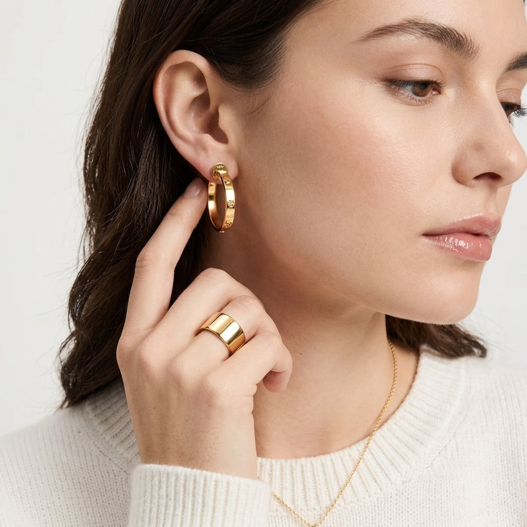 A close-up lifestyle shot of a woman wearing a polished gold hoop earring and a matching gold ring, styled with a delicate gold necklace and a white ribbed knit sweater against a clean, light background.