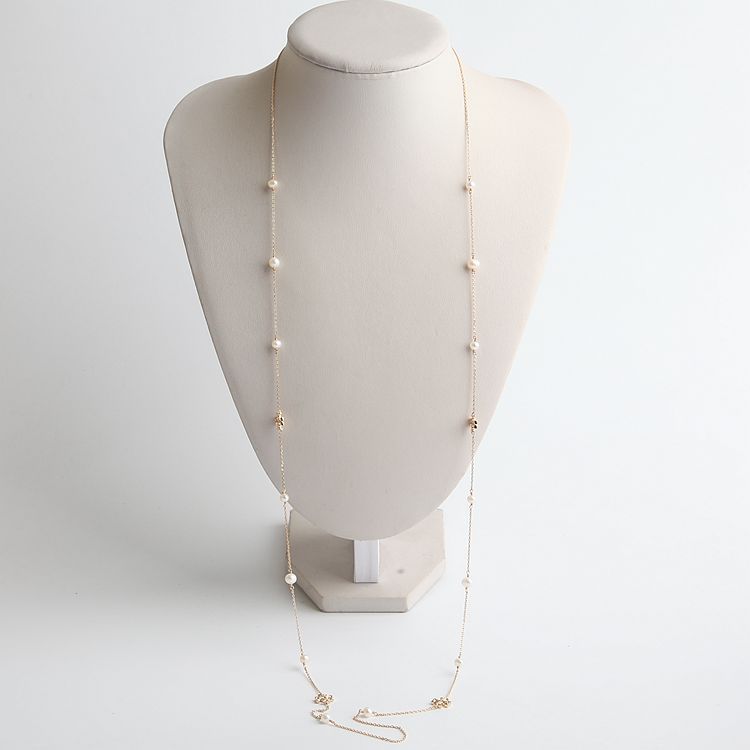 A delicate gold chain necklace with small white pearl-like beads spaced along the chain, displayed on a cream-colored jewelry bust against a light gray background.