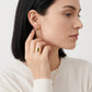 A close-up of a woman wearing a delicate gold hoop earring with a circular medallion design, along with a gold ring and necklace, against a clean white background.