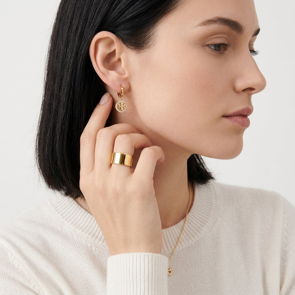 A close-up of a woman wearing a delicate gold hoop earring with a circular medallion design, along with a gold ring and necklace, against a clean white background.