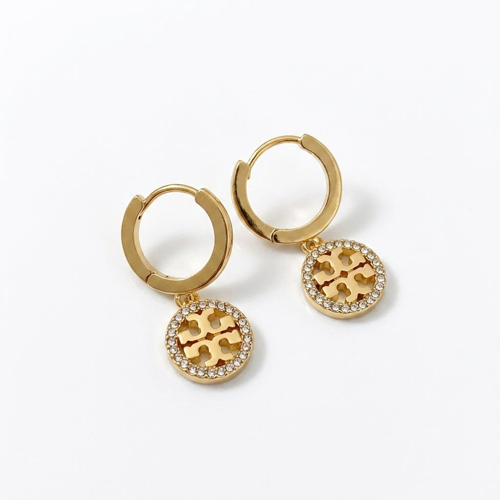 A pair of gold hoop earrings with circular drop charms featuring a raised gold monogram motif surrounded by small clear stones, shown on a clean white background.
