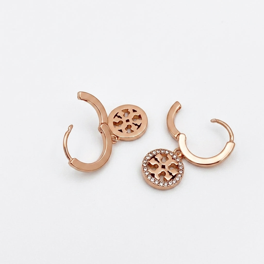 A pair of rose gold huggie hoop earrings with circular medallion charms, one featuring a cutout geometric pattern and the other encrusted with small clear rhinestones, displayed on a clean white background.