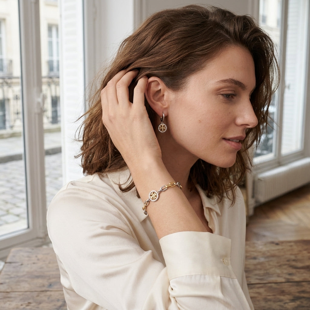 A woman in a cream-colored blouse is shown in profile, wearing a delicate gold chain bracelet and matching drop earrings with circular star-like charms, set against a softly blurred interior background with large windows.