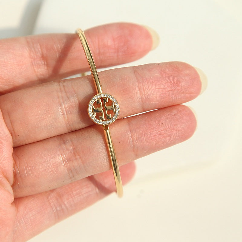 A gold bangle bracelet with a circular charm featuring a four-leaf clover motif surrounded by small clear stones, held in a person's hand against a bright off-white background.