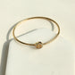 A gold bangle bracelet with a central geometric logo charm, displayed on a white jewelry stand.