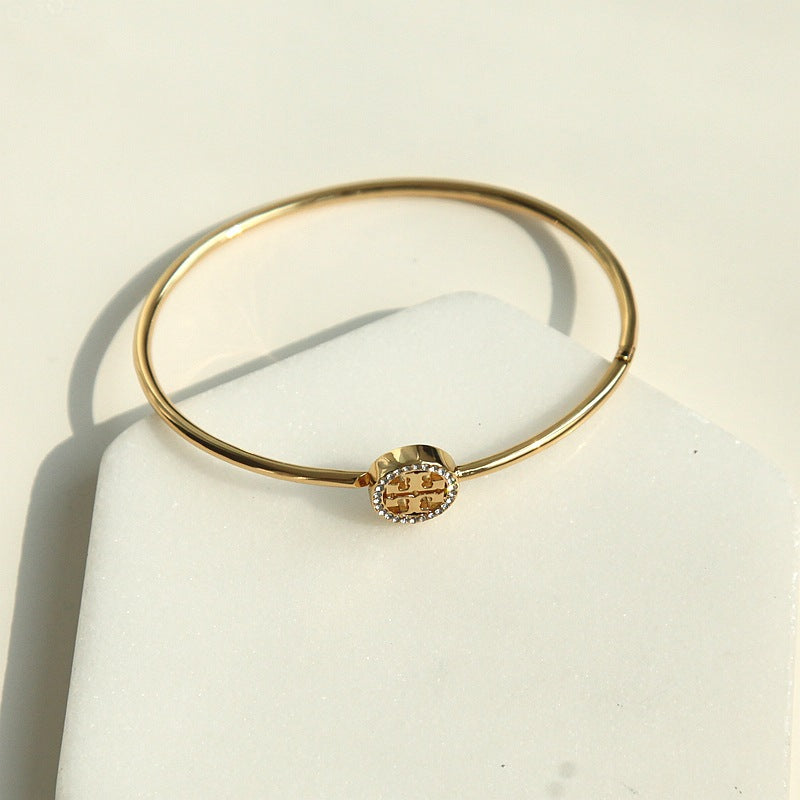 A gold bangle bracelet with a central geometric logo charm, displayed on a white jewelry stand.