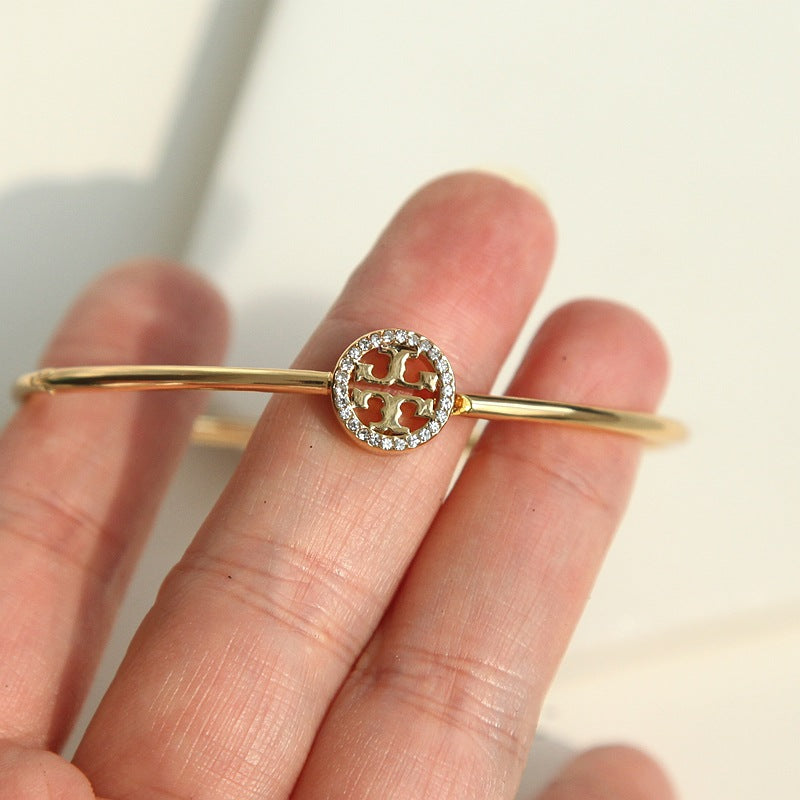 A gold bangle bracelet with a circular logo charm, held on a person's fingers. The charm features a four-leaf clover design surrounded by small clear stones.