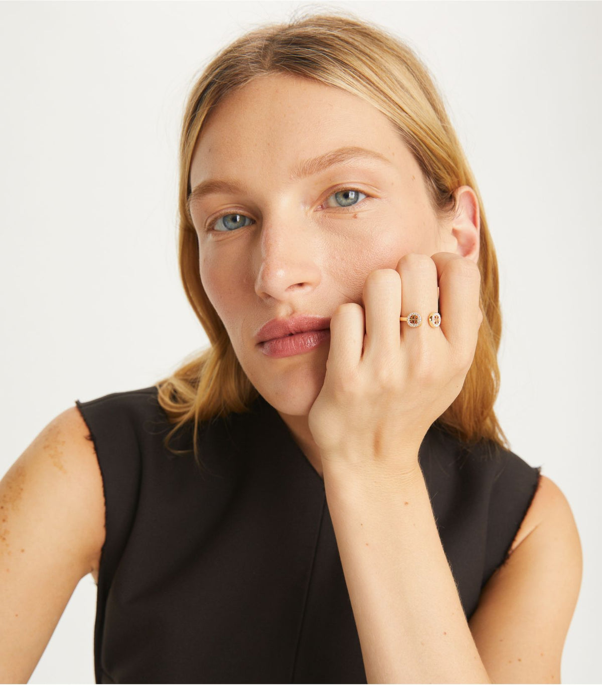 A close-up portrait of a woman with blonde hair wearing a black sleeveless top and delicate gold rings on her fingers, set against a clean white background.