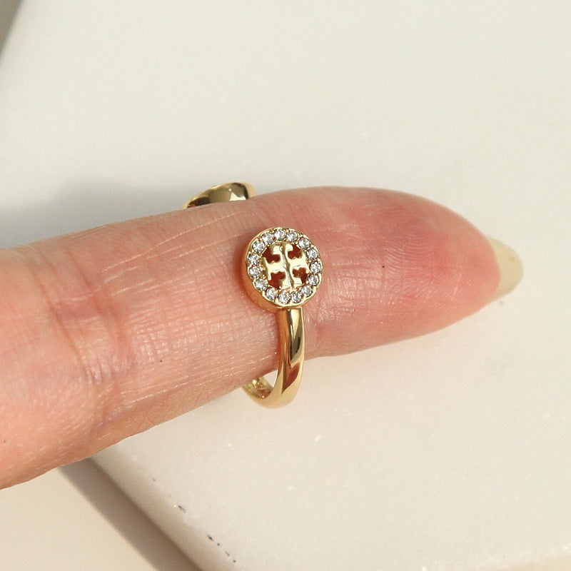 A gold ring with a round crystal halo and a central red cross motif, shown worn on a finger against a light off-white background.