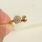 A close-up of a hand holding a gold ring with a round halo of small clear stones and a textured center, shown against a light neutral background.