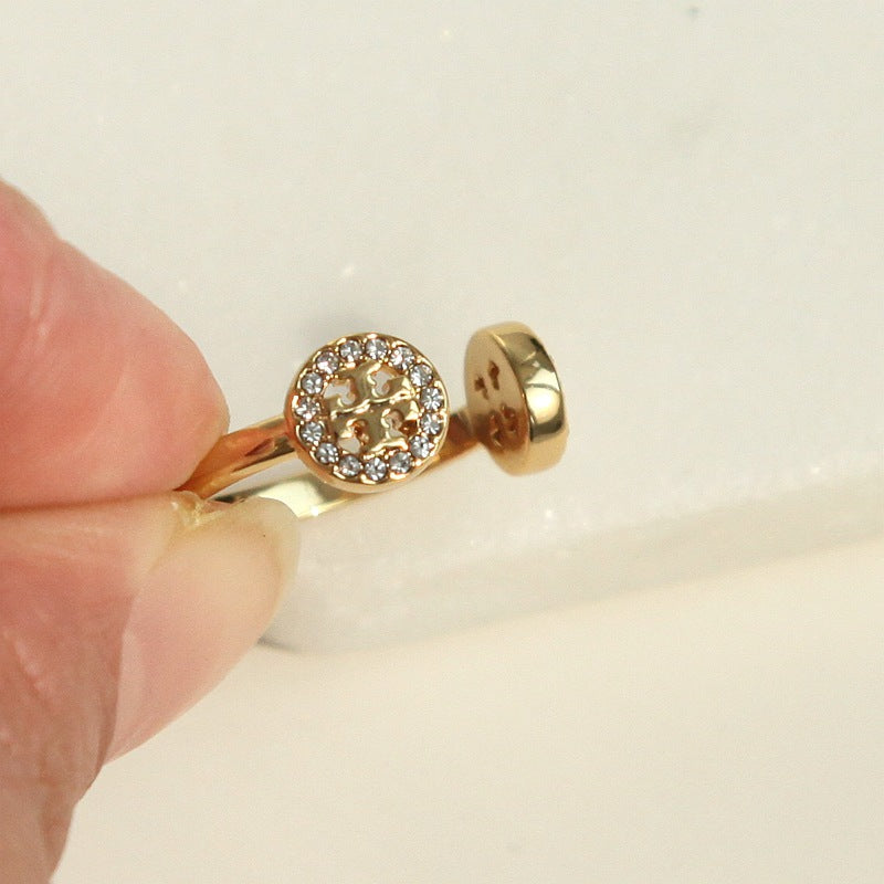 A close-up of a hand holding a gold ring with a round halo of small clear stones and a textured center, shown against a light neutral background.