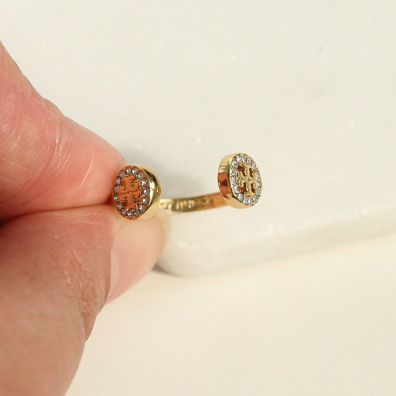 A close-up of a hand holding a gold ring with two oval pavé-set stones, one featuring a brown monogram and the other a silver monogram, on a white background.