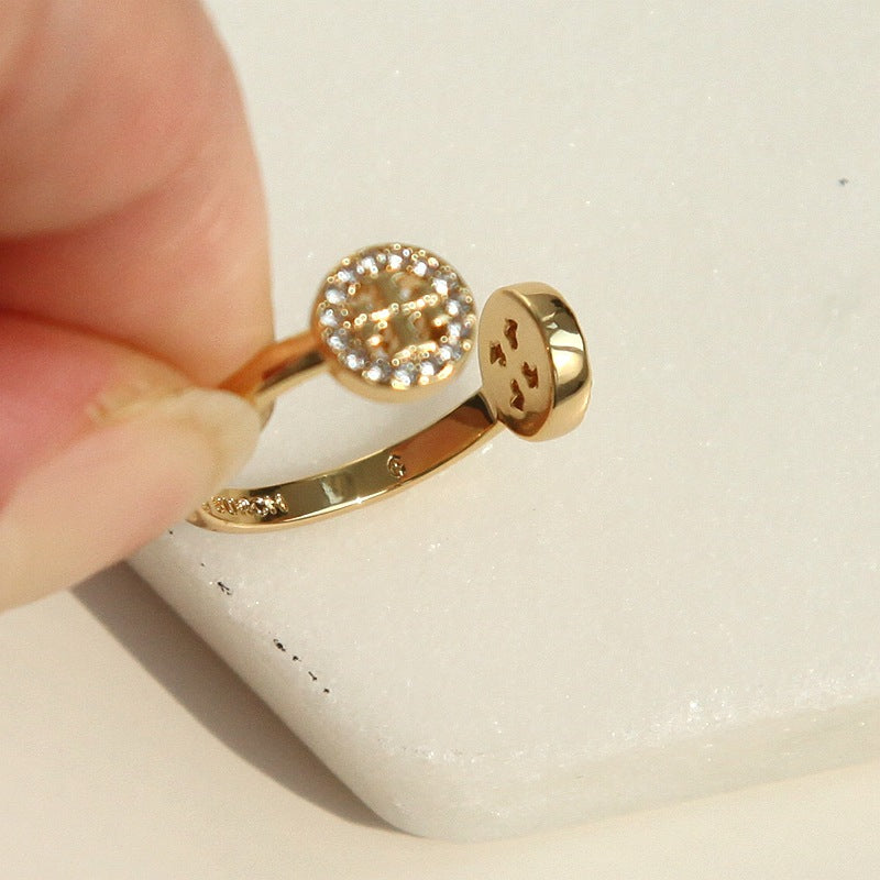 A gold ring with a round pavé-set crystal top and a flat oval side detail, held by a hand against a white textured background.