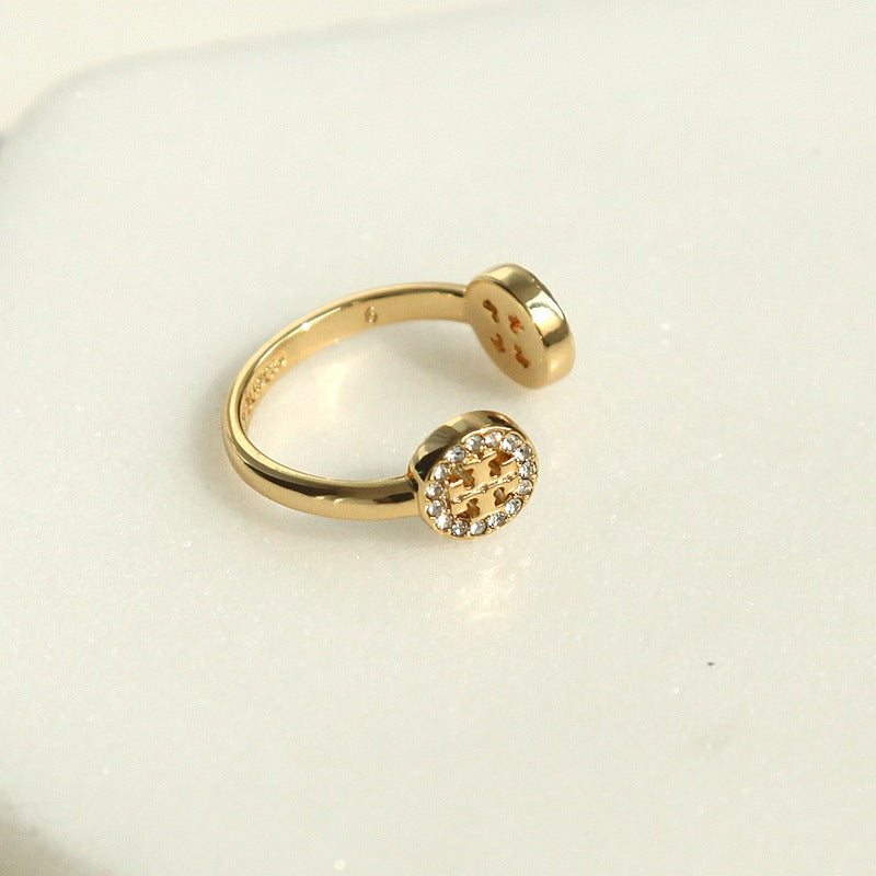 A gold open-ended ring featuring a round bezel-set cluster of small clear stones with a central gold accent, displayed on a clean white surface.