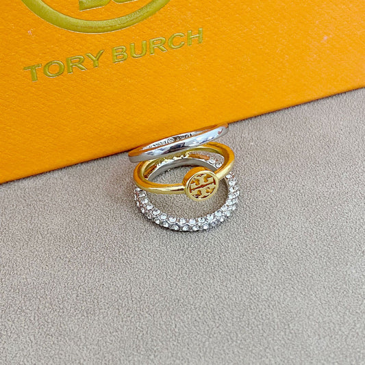 Two rings, one gold and one silver with a central logo, on a gray surface with a Tory Burch box in the background.