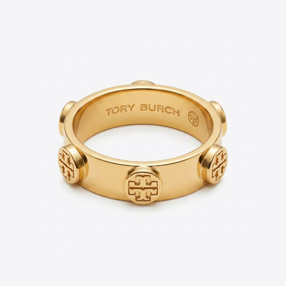 A gold-tone Tory Burch ring with a wide band, featuring a central embossed monogram logo and three matching monogram details spaced around the band, shown on a light gray background.