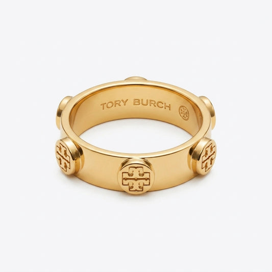 A gold-tone Tory Burch ring with a wide band, featuring a central embossed monogram logo and three matching monogram details spaced around the band, shown on a light gray background.