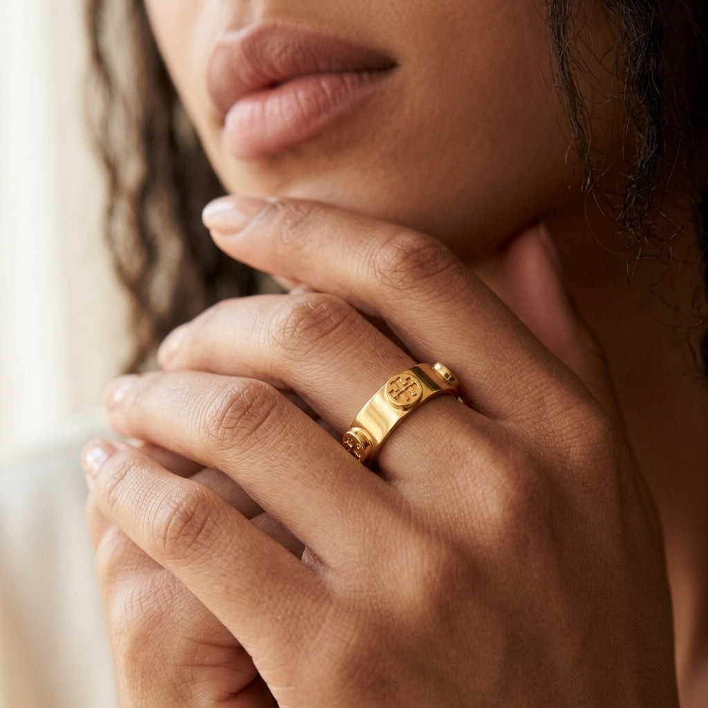 A close-up lifestyle shot of a model wearing a gold signet ring with an engraved circular emblem on the ring finger.