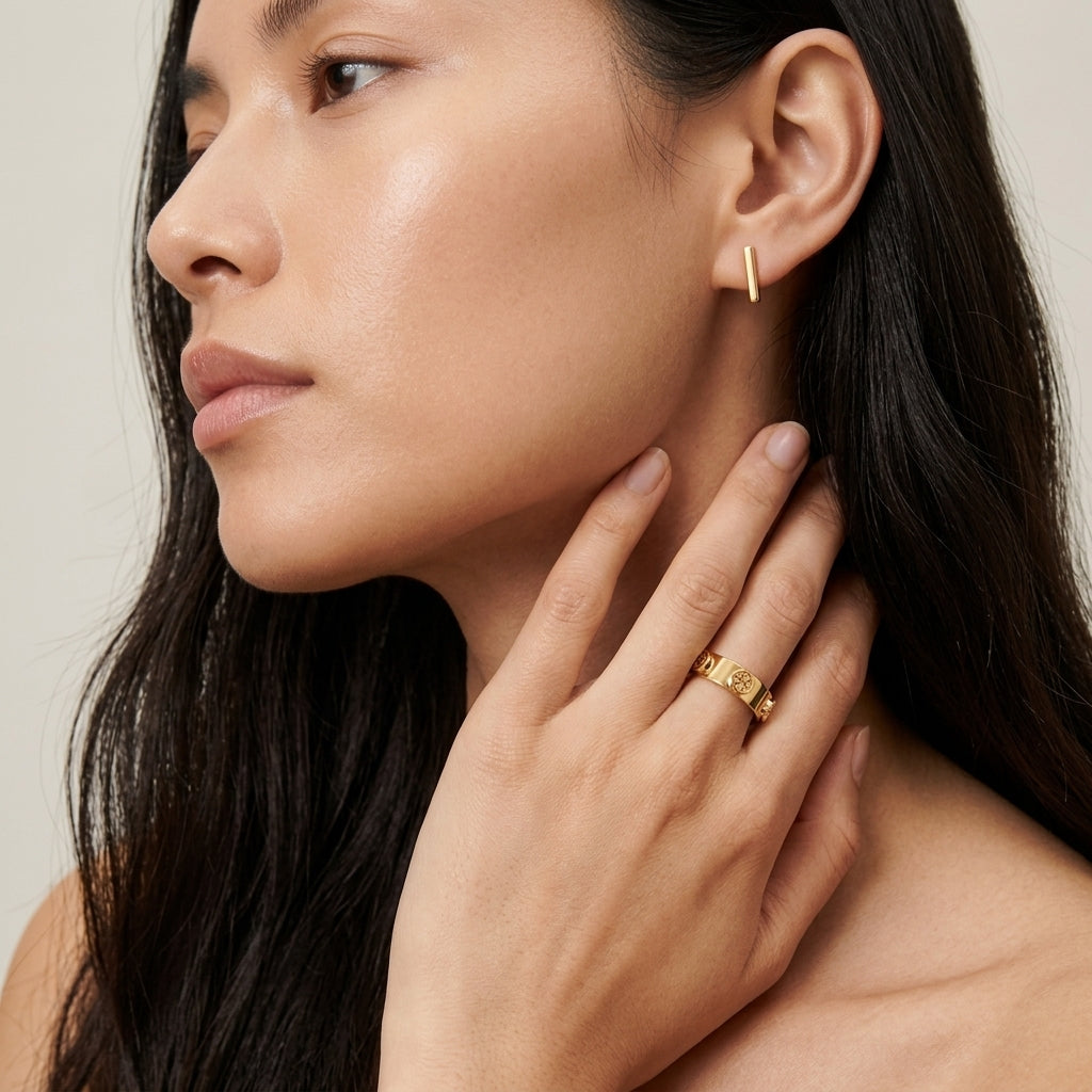 A close-up of a woman wearing a delicate gold ring on her finger and a small gold hoop earring, with her hand resting near her neck against a soft neutral background.
