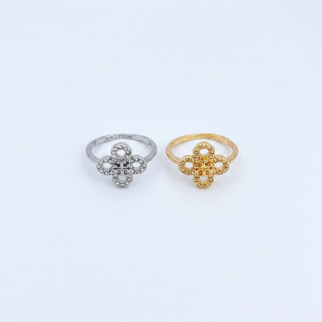 Two small flower-shaped rings on a plain white background, one in silver and one in yellow gold. The silver ring has clear stones, and the gold ring has textured openwork detailing.