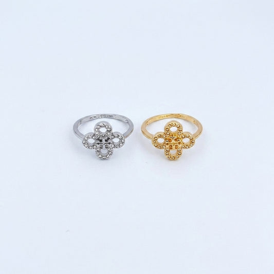 Two small flower-shaped rings on a plain white background, one in silver and one in yellow gold. The silver ring has clear stones, and the gold ring has textured openwork detailing.