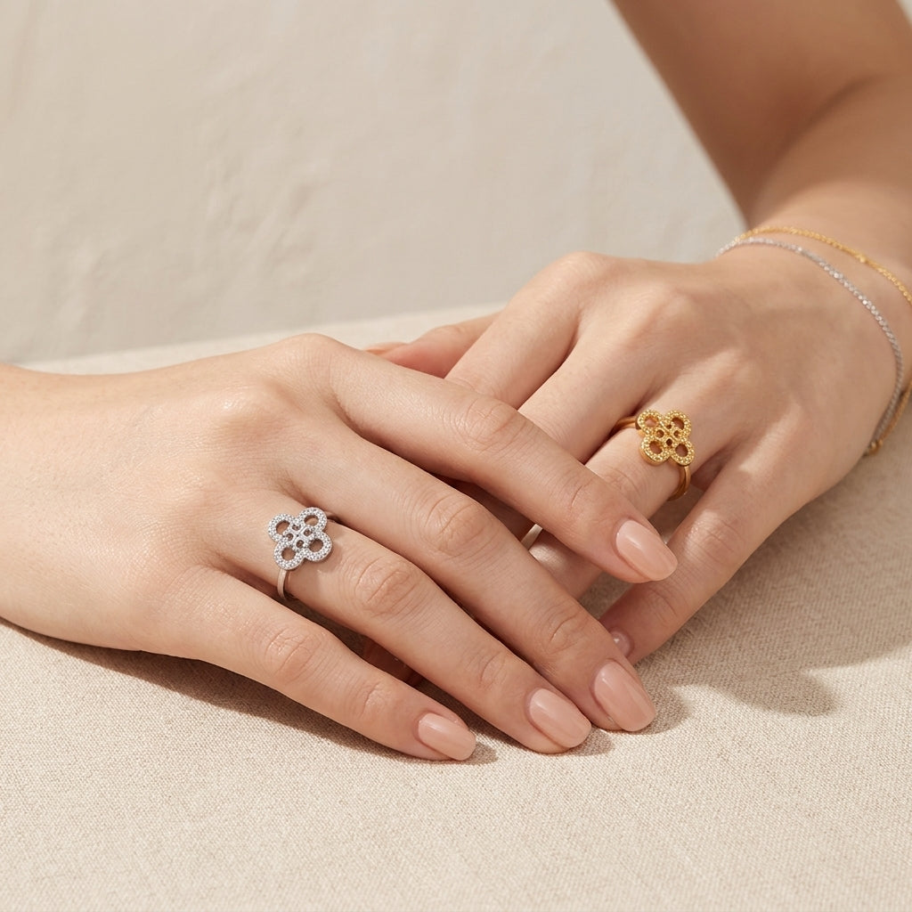 Two hands resting on a beige fabric surface, each wearing a small flower-shaped ring. One ring is silver with clear stones, and the other is gold.