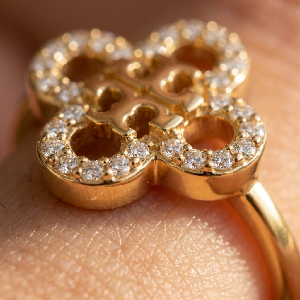 A close-up of a gold ring with a clover-like four-petal design, set with small clear stones around the edges, worn on a finger.
