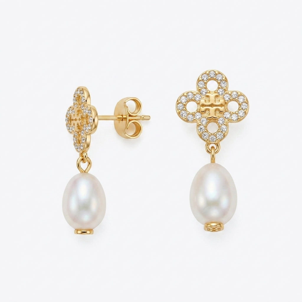 A pair of gold dangle earrings featuring a pavé-set floral clover motif with a small central logo and a dangling freshwater pearl, shown on a light gray background.