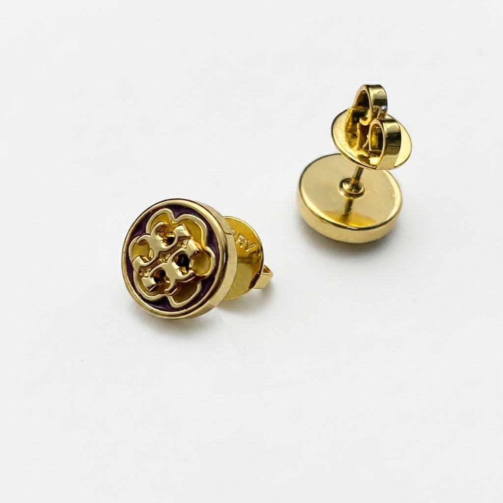 A pair of gold stud earrings on a white background, with one earring showing the front face featuring a raised quatrefoil logo and the other showing the side profile with the post and backing.