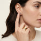 A close-up of a woman wearing gold jewelry, featuring a small gold hoop earring, a gold stud earring with a circular logo, and a wide gold ring on her finger.
