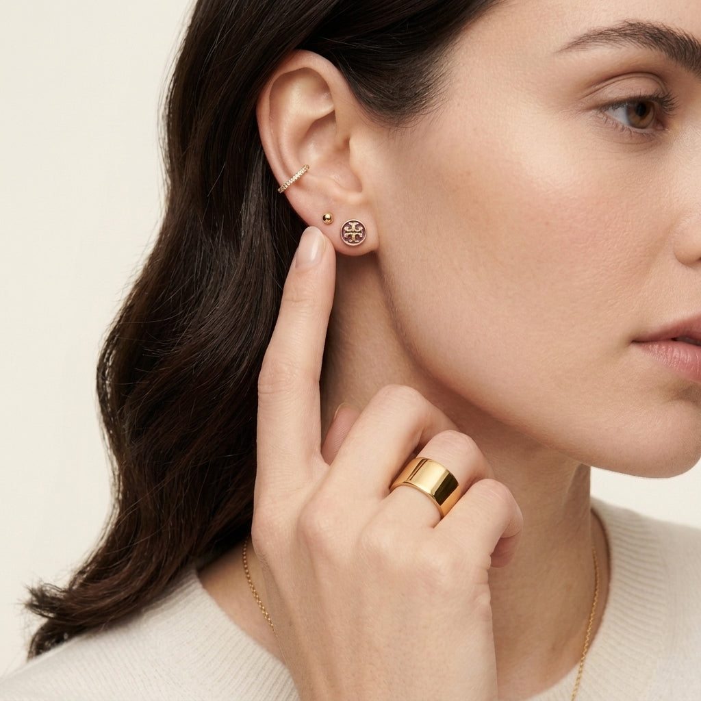 A close-up of a woman wearing gold jewelry, featuring a small gold hoop earring, a gold stud earring with a circular logo, and a wide gold ring on her finger.