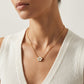 A close-up of a model wearing a delicate gold chain necklace with a small white flower-shaped pendant featuring a gold center, styled with a white ribbed knit top.